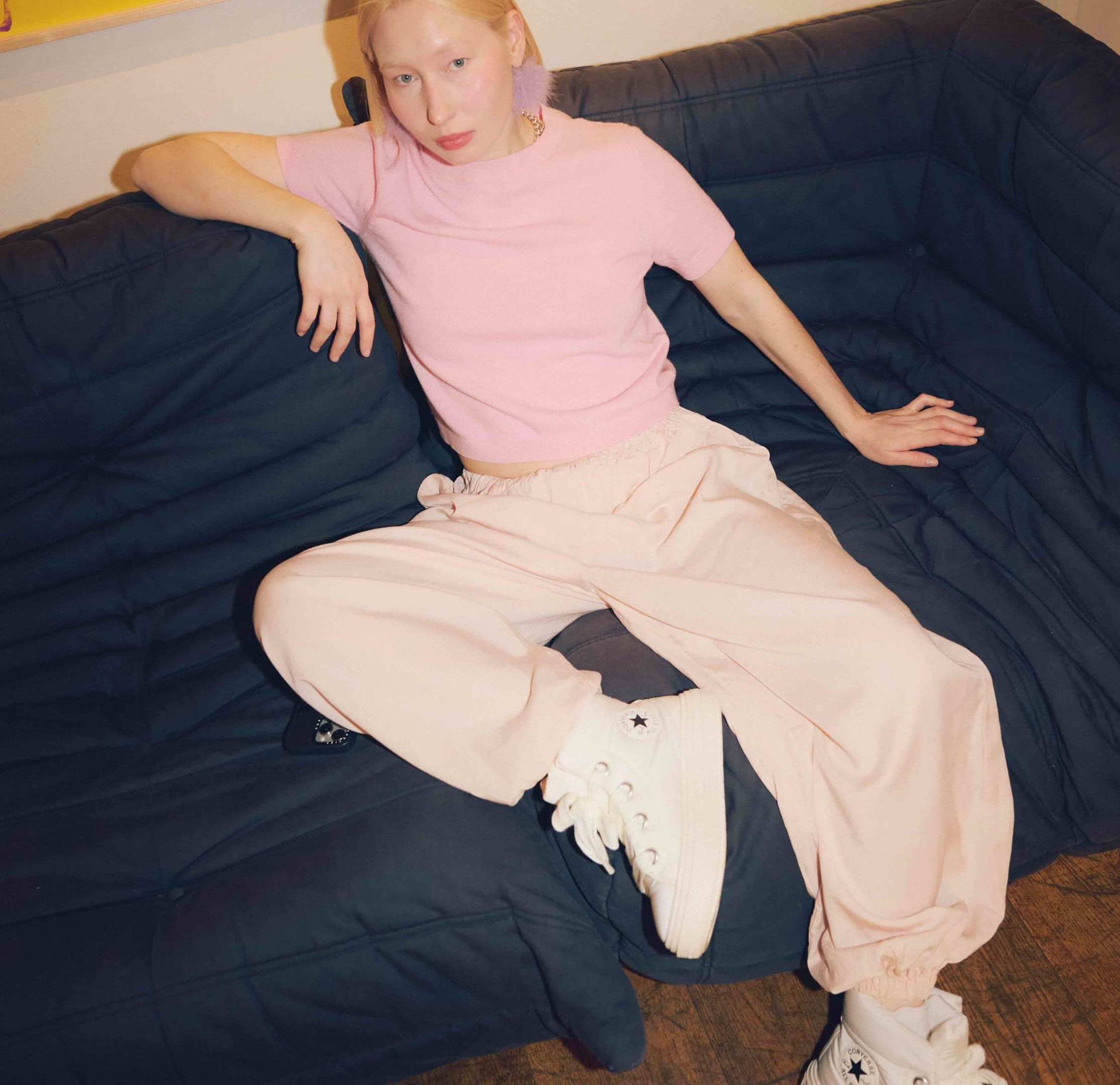 Top de manga corta color rosa claro combinado con pantalones sueltos color rosa pálido y zapatillas blancas de caña alta. Sentado en un sofá negro acolchado.