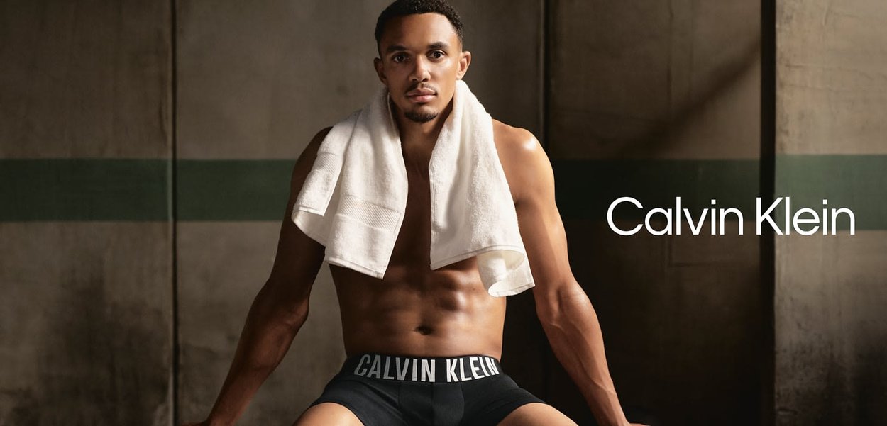 Schwarze Calvin Klein Boxershorts mit weißem Logo-Taillengummi; Model mit kurzem Haar, weißem Handtuch über den Schultern, sitzend an einer Wand.