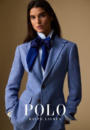 Une femme porte un blazer bleu clair et une chemise blanche avec un grand foulard nœud bleu marine, se tenant devant un fond beige, les mains dans les poches.