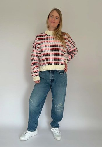 Vit tröja med röd och svart randig mönster, i kombination med ljusblå ripped jeans och vita sneakers, stående mot en neutral bakgrund.