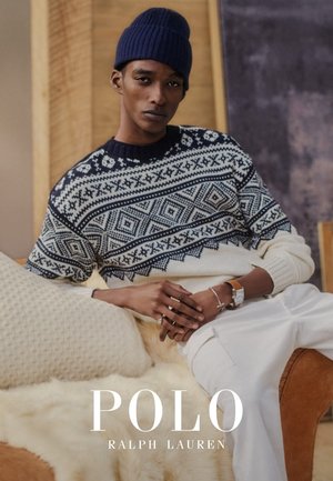 Man met blauwe gebreide muts, een marineblauw-witte trui met patroon, witte broek, bruine horloge, zittend op een beige bank met gestructureerde kussens.