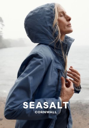 Kvinde i blå regnjakke med hætte op står tæt på vandet, øjnene lukkede, hænderne nær brystet, fremstår rolig og reflekterende udendørs.