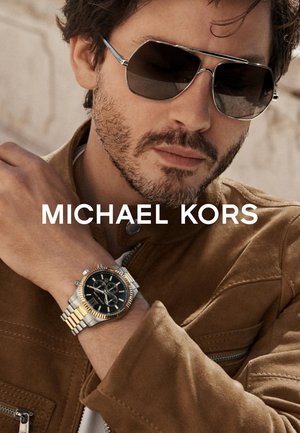 Giacca in suede marrone, orologio in metallo argento e oro con quadrante nero e dettagli cronografici, occhiali da sole scuri con accenti blu.