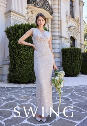 Frau in einem ärmellosen, bodenlangen weißen Spitzenkleid, die einen Blumenstrauß hält und auf einem Steinpflaster vor einem kunstvollen Gebäude mit Grün steht.