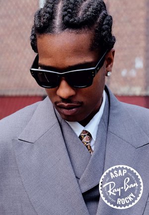 Man met gevlochten haar draagt zwarte Ray-Ban zonnebril, grijs pak, wit shirt, gestreepte stropdas en diamantoorbel, kijkend naar beneden.