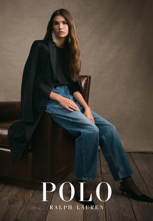 Blazer noir sur un top noir, jean bleu taille haute et sandales à talons. Boucles d'oreilles en perles grandes. Arrière-plan chaud et neutre, sol en bois.