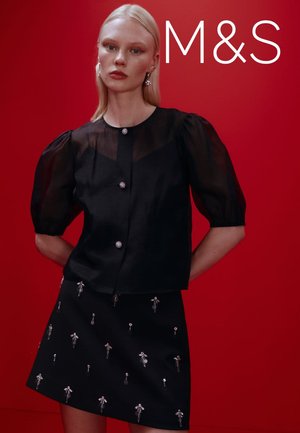 Czarna koszula zapinana na guziki z przezroczystymi, bufkami na rękawach, zestawiona z czarną mini spódniczką zdobioną metaliczną, kwiatową haftą. Tło w kolorze czerwonym.