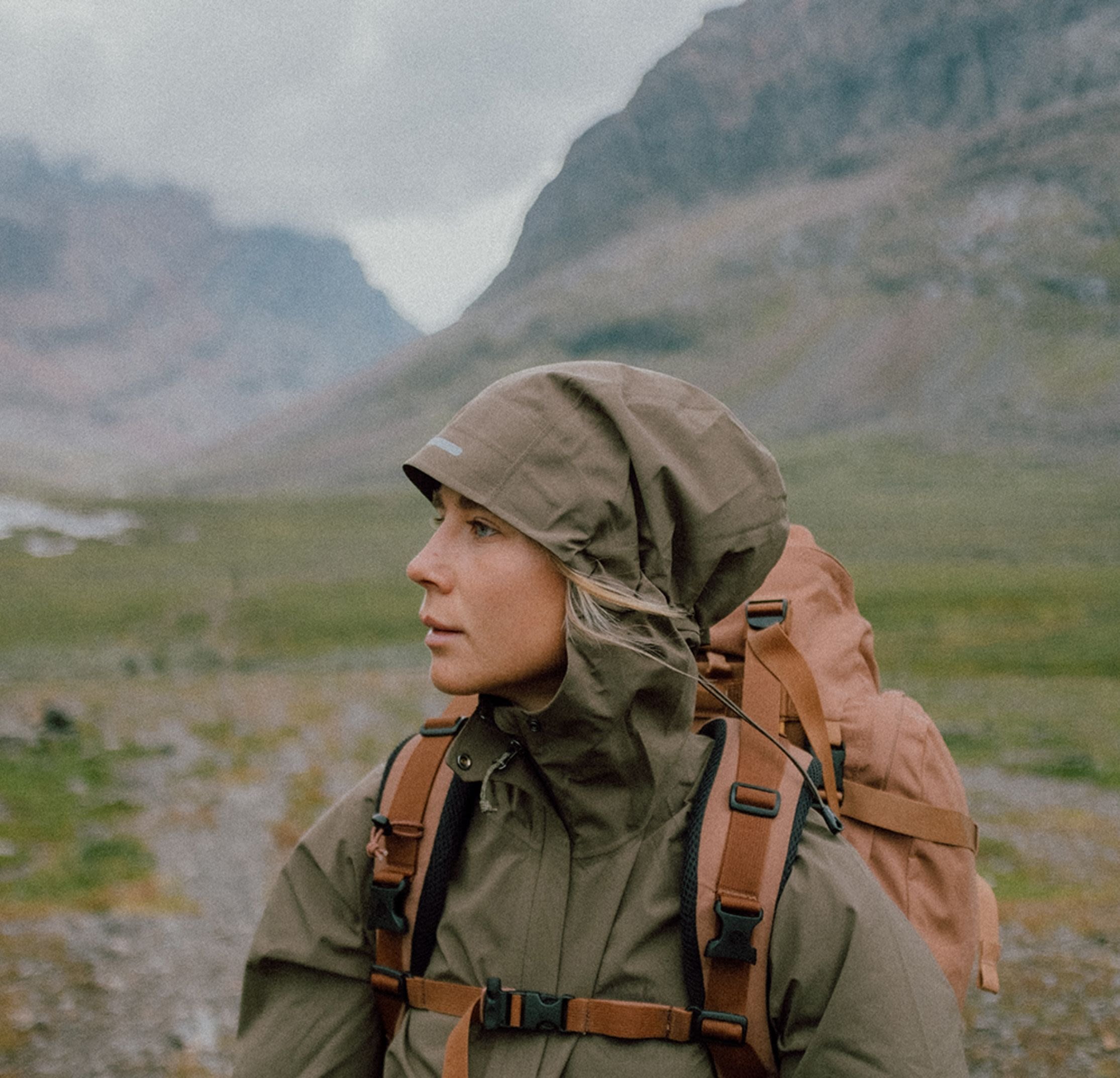 Brun vattentät jacka med luva, justerbar dragsko, och tan ryggsäck med bruna remmar, placerad mot en bergig landskap.