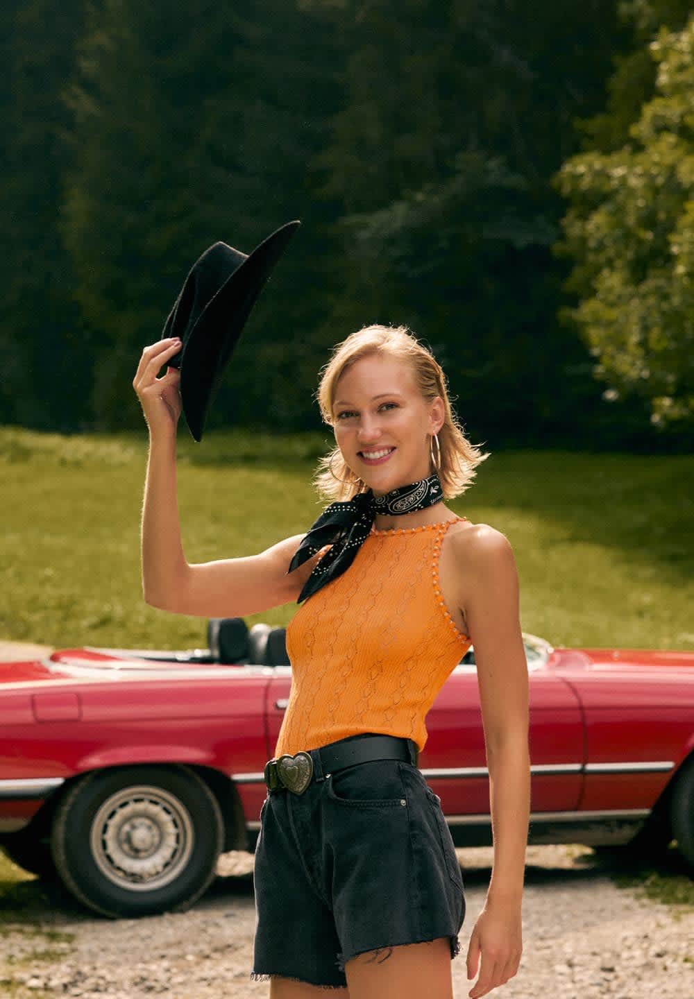 Un débardeur en tricot orange, un short en denim noir et un bandana noir ; se tenant à côté d'une décapotable rouge avec un arrière-plan vert.