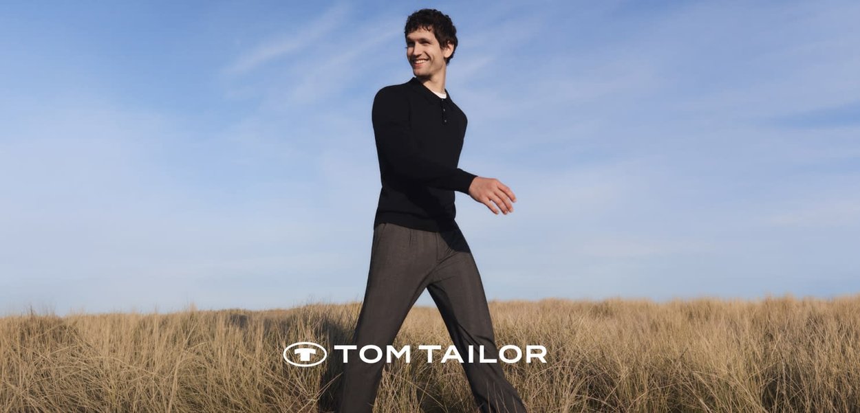 Mann in schwarzem Pullover und grauer Hose, der durch trockenes Gras unter blauem Himmel läuft, lächelt und zur Seite schaut