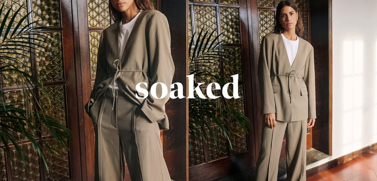 Hellolivgrüner, maßgeschneiderter Anzug mit Wickeldesign, ausgestattet mit Kordelzug in der Taille, Taschen und weichem Stoff. Im Hintergrund sind Holzpaneele zu sehen.