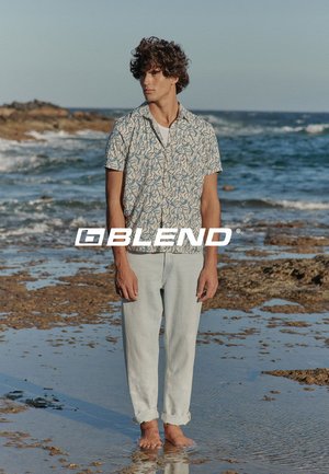 Young man barefoot stands on rocky shore wearing light patterned short-sleeve shirt and rolled-up light jeans with ocean waves in the background.