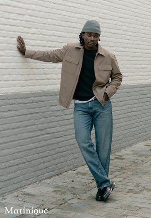 Man in gray beanie, beige jacket, black shirt, and blue jeans leans against white brick wall with one hand, standing on cobblestone pavement.