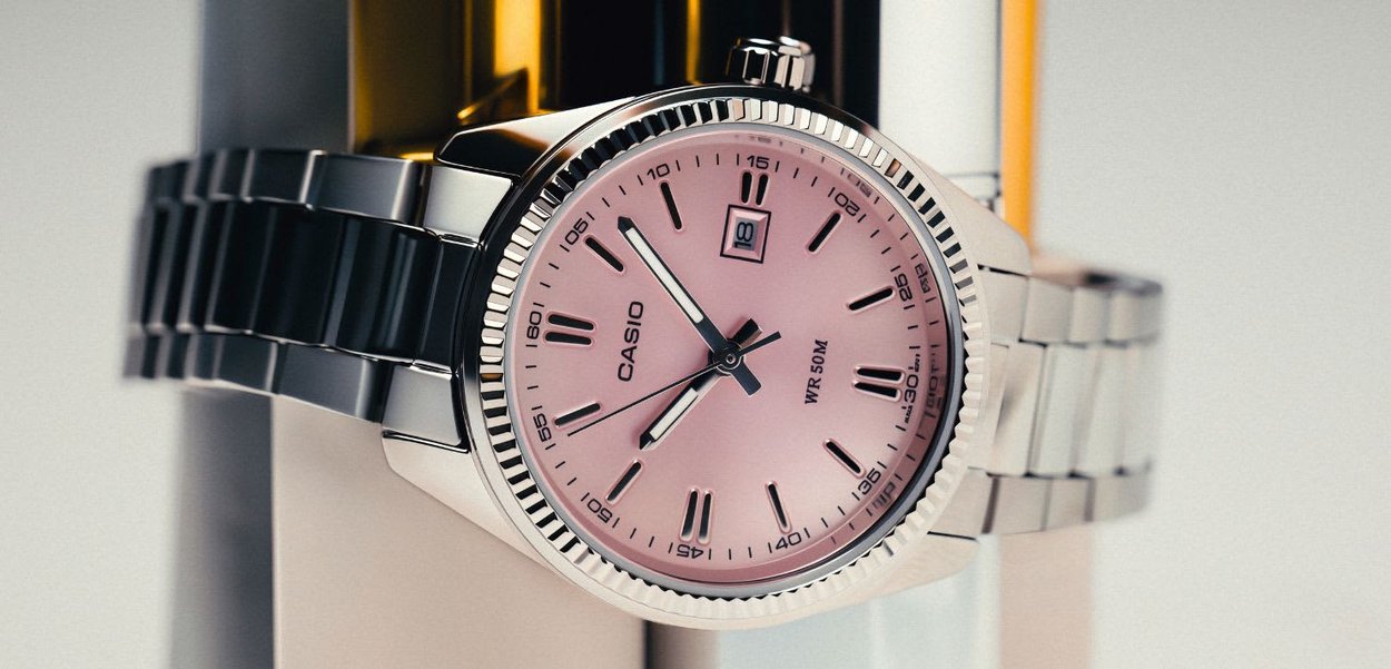 Reloj de pulsera Casio plateado con esfera rosa, marcadores de hora negros, pantalla de fecha y correa de eslabones metálicos sobre un fondo desenfocado.