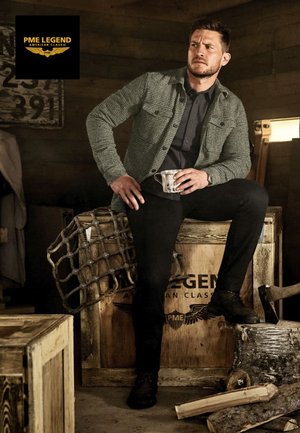 Green textured jacket over a dark shirt, black pants, and black shoes. Holding a metal cup, standing on a wooden box in a rustic setting.