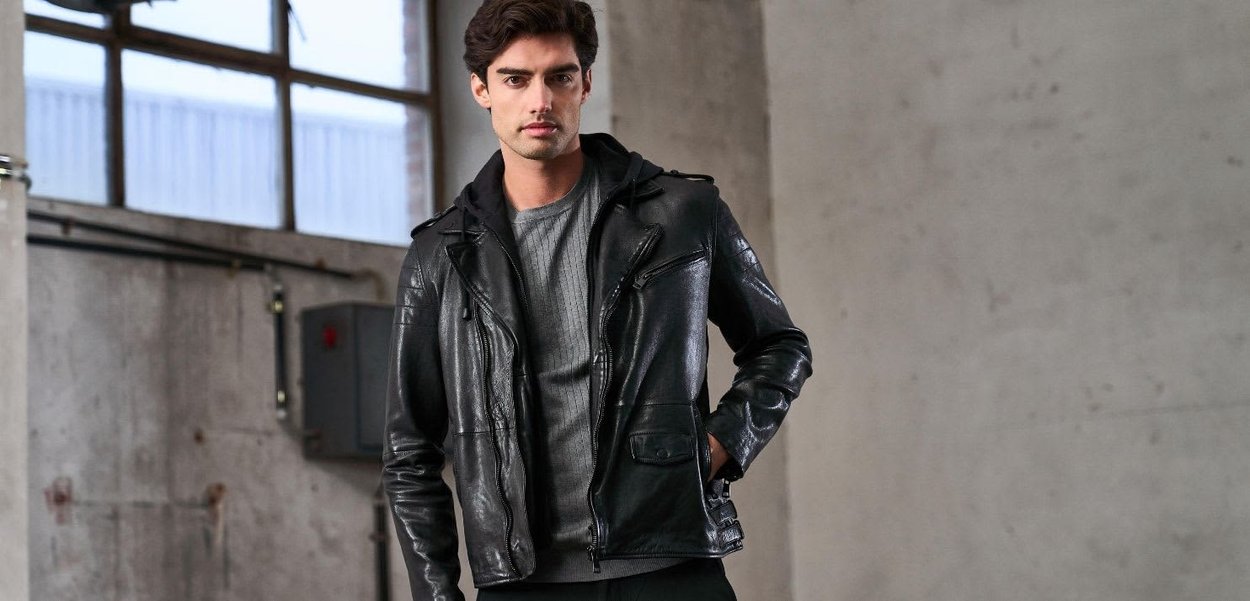 Giacca da motociclista in pelle nera con tasche con cerniera, abbinata a un maglione grigio a coste e un cappuccio nero. Texture liscia, design aderente.