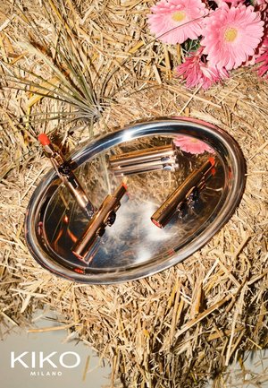 Zwei Lippenstiftbehälter in roségoldener Metallhülle mit roten Spitzen liegen auf einem silbernen Tablett, umgeben von Stroh und pinken Blumen.