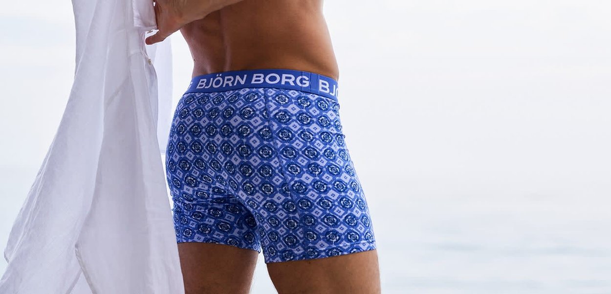 Mand iført blå mønstrede Björn Borg boxerbriefs, der holder en hvid skjorte, med en klar himmel i baggrunden.