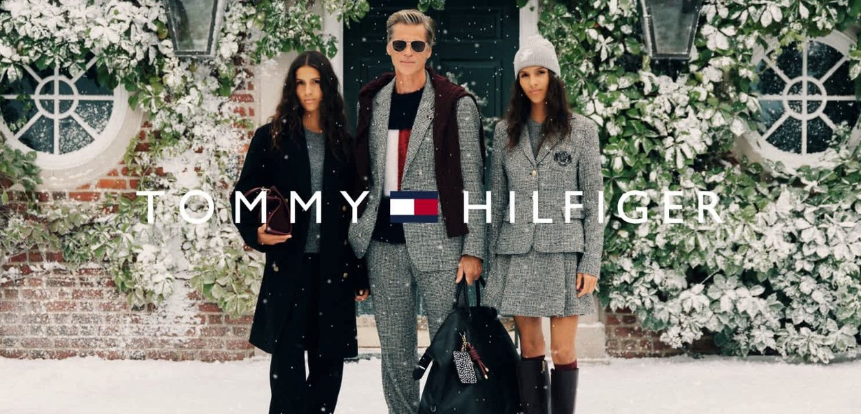 Tre persone in abbigliamento invernale sono in piedi davanti a uno sfondo innevato con vegetazione verde. La prima indossa un lungo cappotto nero, la seconda un completo grigio e la terza un blazer grigio con motivo a quadri.