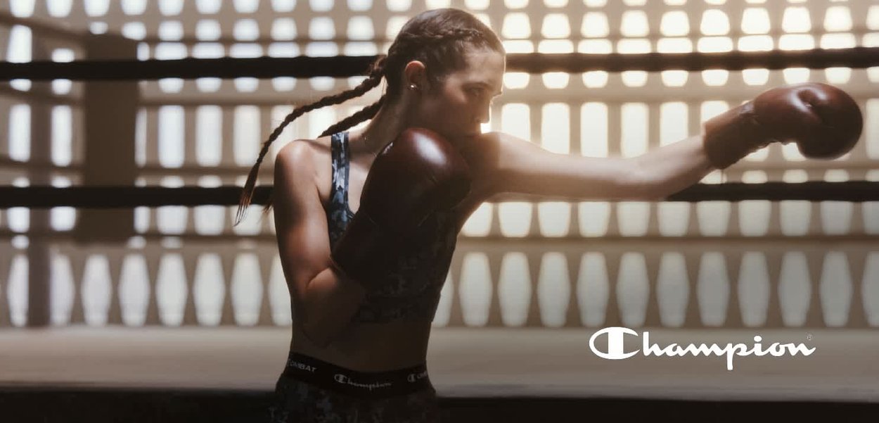Boxeadora con guantes marrones y un sujetador deportivo negro con patrón, lanzando un golpe en un gimnasio bien iluminado con patrones de fondo geométricos.