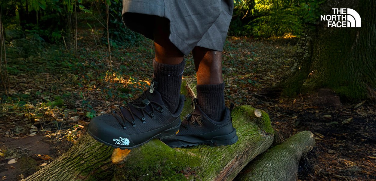 Chaussures de randonnée noires avec un mesh texturé et des semelles en caoutchouc, portant des chaussettes grises, posées sur une bûche dans une zone boisée.