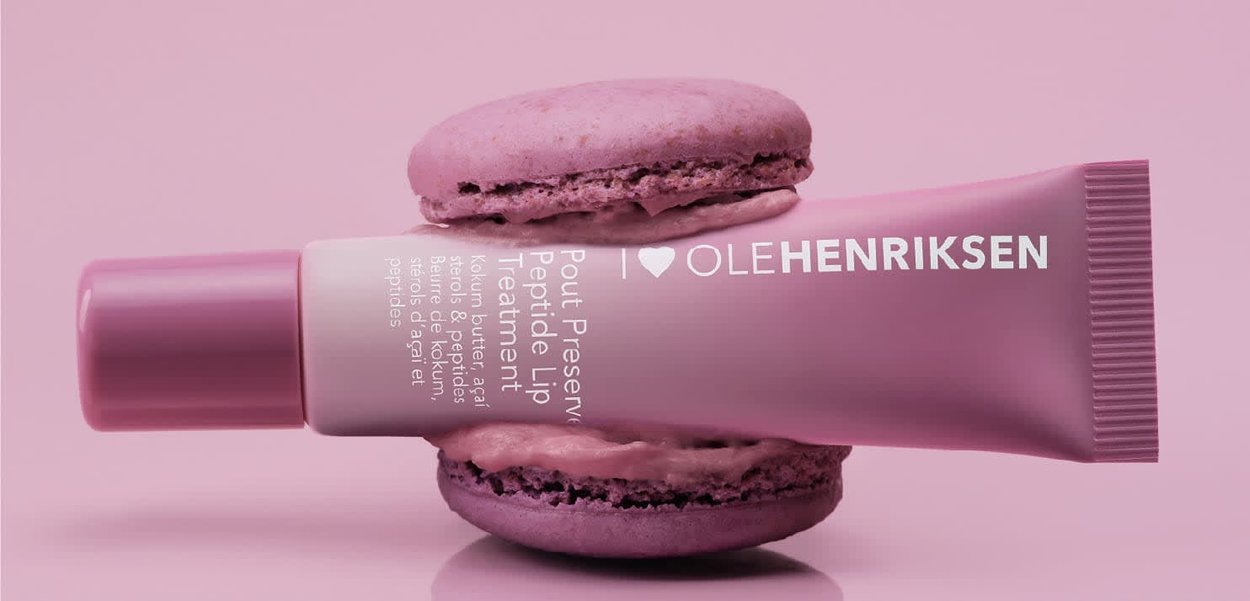 Pinkfarbige Lippenpflege von Ole Henriksen, horizontal platziert zwischen zwei passenden pinken Macarons vor einem sanftrosafarbenen Hintergrund.