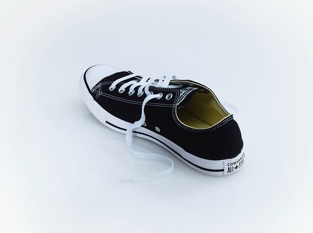 Black canvas sneaker with white rubber sole and toe cap. Features white laces and yellow interior lining. "Converse All Star" branding on heel.