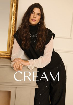 Schwarzes, gestricktes ärmelloses Oberteil mit hohem Kragen und Perlenverzierungen, über einer weißen, durchsichtigen Langarmbluse getragen, kombiniert mit schwarzen Hosen.