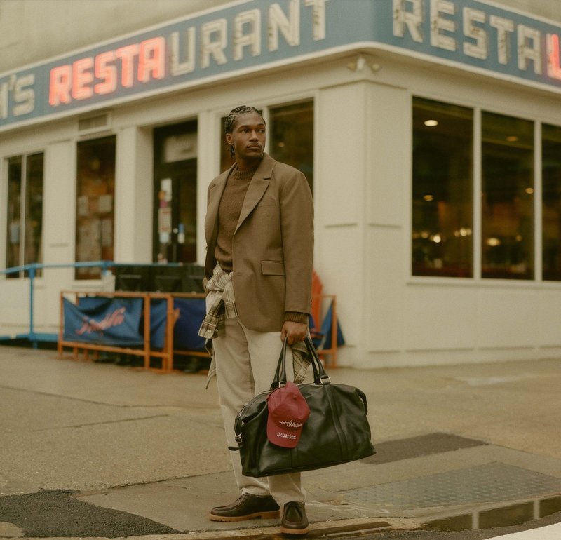 Brun blazer over en beige sweater, lyse bukser, sort læder duffel taske, rød kasket, står udenfor en restaurant.