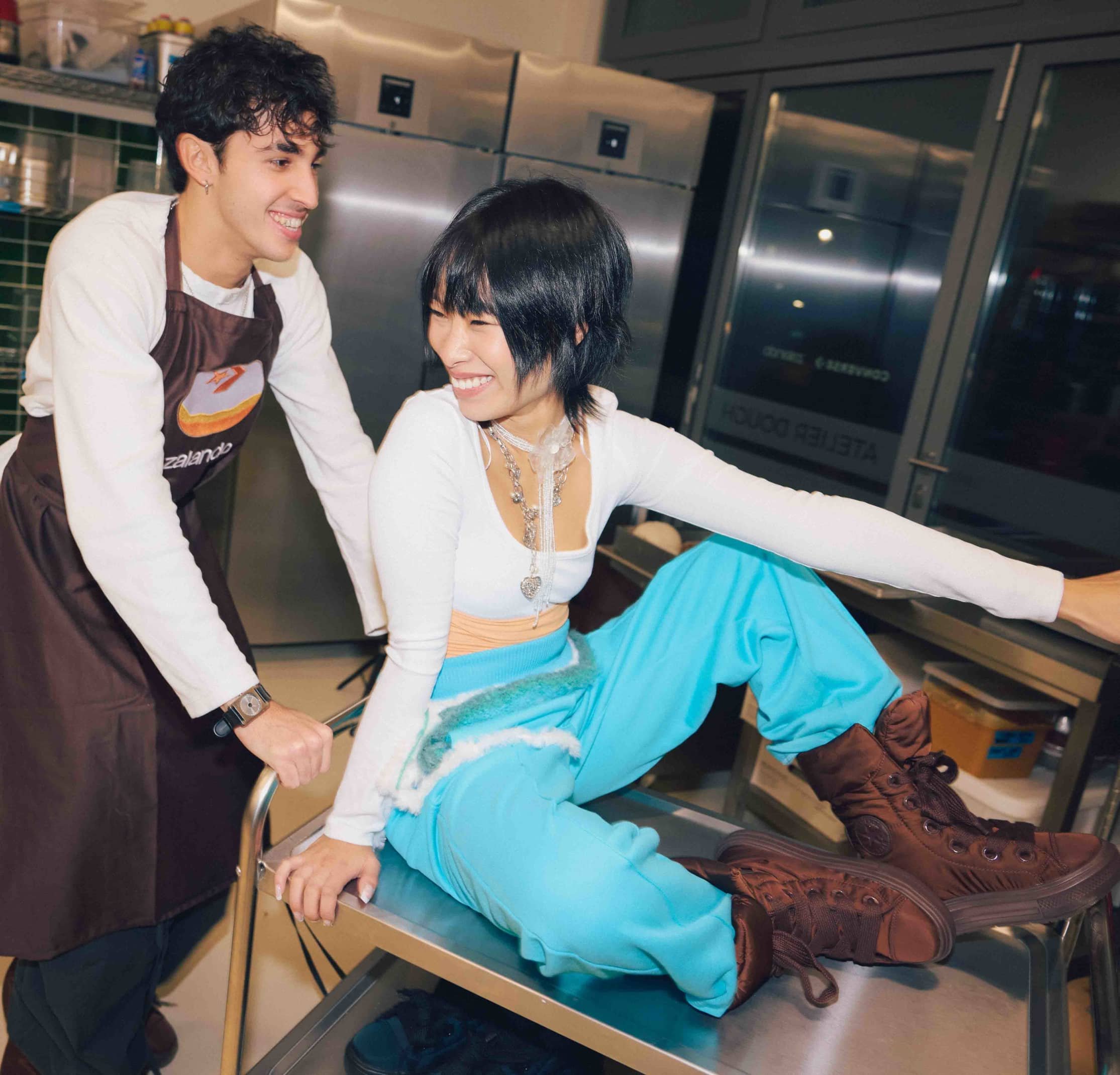 Dos personas en una cocina. Una lleva un delantal marrón y la otra viste una blusa blanca, pantalones azules y botas marrones altas.