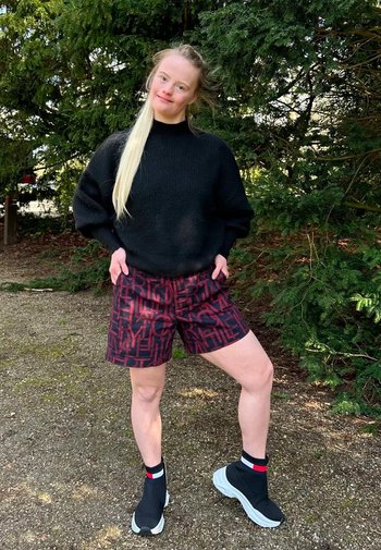 Svart texturerad tröja, mönstrade bordeauxfärgade shorts och svarta ankelstövlar med röda och vita detaljer. Står utomhus med grönska i bakgrunden.