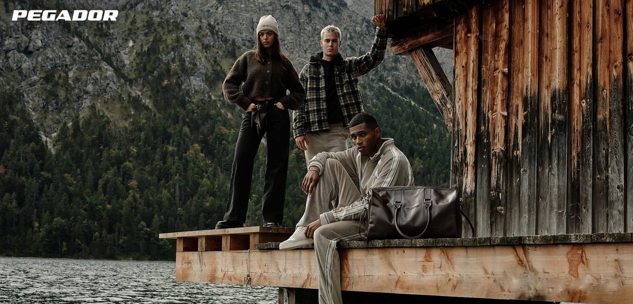 Tre modelli posano su un molo di legno, indossando abbigliamento casual: cardigan marrone testurizzato, giacca a quadri e tuta leggera. Vicino si trova una borsa scura in pelle.