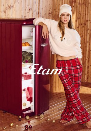 Rode vintage koelkast met open deur, gevuld met fruit en ingepakte cadeaus. Model draagt een crèmekleurige trui en rode geruite broek voor een houten achtergrond.