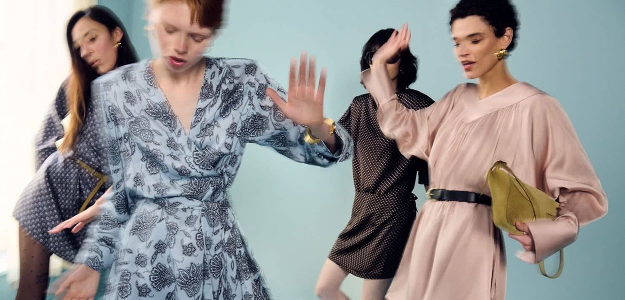 Vier Frauen in gemusterten und einfarbigen Kleidern, die vor einem schlichten hellblauen Hintergrund tanzen oder sich ausdrucksstark bewegen.