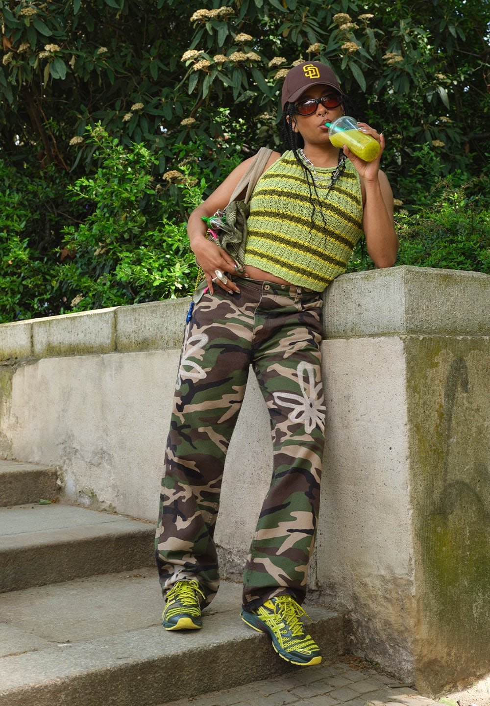 Verde e amarelo às riscas, top de malha, calças de camuflagem com padrões florais brancos, boné preto, óculos de sol e ténis neon. Segurando uma bebida.