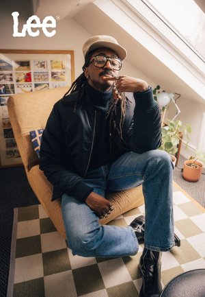Schwarze Bomberjacke über einem dunklen Rollkragenpullover, lockere blaue Denim-Jeans und glänzende schwarze Knöchelstiefel. Trägt eine Brille und eine helle Mütze.