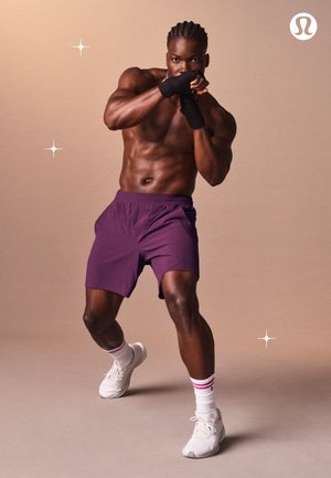 Athletische Shorts aus lila Stoff; muskulöse Figur in Boxhaltung; weiße Sportschuhe; strukturierte Handgelenkbandagen; rosa-weiß gestreifte Socken.