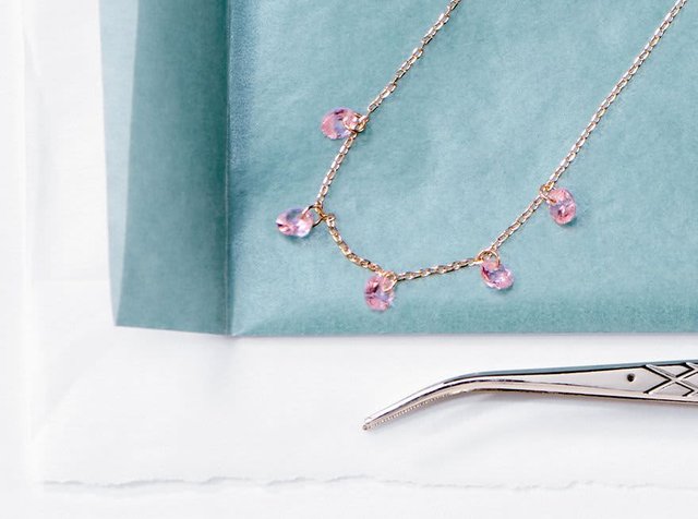 Χρυσή αλυσίδα κολιέ με πέντε μικρούς ροζ σταγόνας γοητείες πάνω σε μπλε ύφασμα, δίπλα σε γωνιακά μεταλλικά τσιμπίδα σε λευκή επιφάνεια.