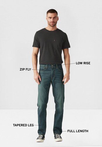 Camiseta de color gris oscuro, vaqueros azul denim con cremallera, tiro bajo, pierna estrecha, largo completo y zapatillas en blanco y negro.