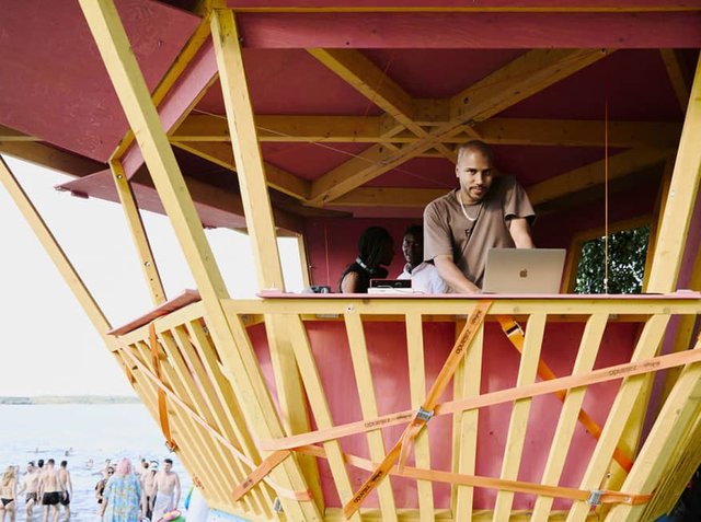 Holz-Rettungsturm mit einem pinken und gelben Design, der über eine offene Struktur, diagonale Verstärkungen und eine Person verfügt, die drinnen einen Laptop benutzt.