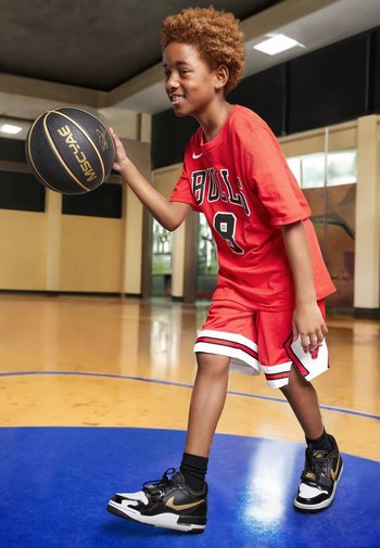 Uniforme de baloncesto rojo con detalles en negro, que incluye camiseta y pantalones cortos. El niño sostiene un balón de baloncesto negro, vistiendo zapatillas negras y doradas en una cancha de madera.