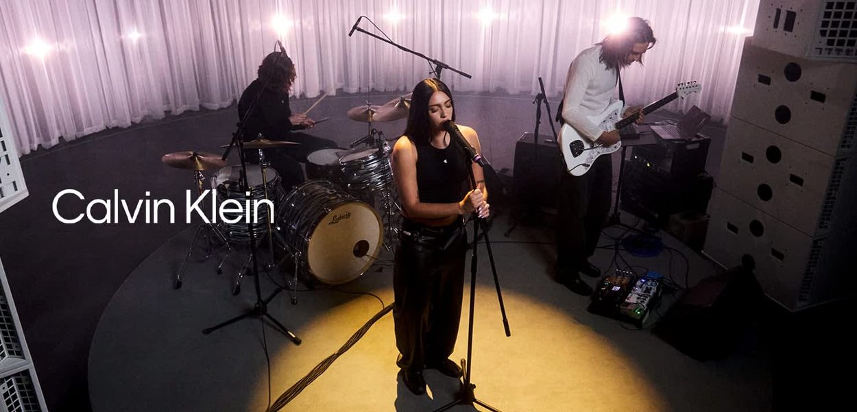 Una cantante actúa frente a un micrófono, con un batería y un guitarrista detrás de ella en un estudio; a la izquierda aparece el texto Calvin Klein.