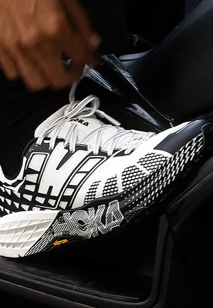Person's hand fastening a white and black Hoka running shoe while seated inside a vehicle.