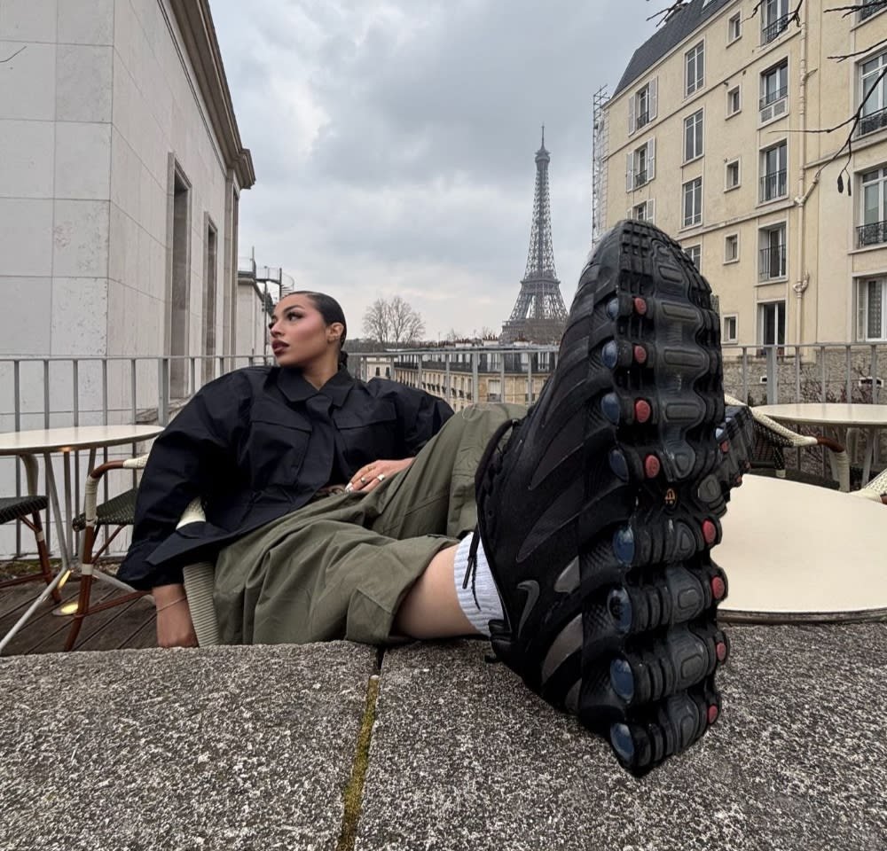 Black athletic shoes with textured soles, red and blue accents; model wearing a loose black jacket and olive green pants, Eiffel Tower in background.