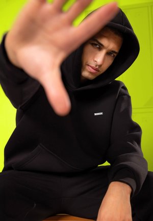 Young man in black hoodie sitting, reaching hand toward the camera against a bright yellow-green background.