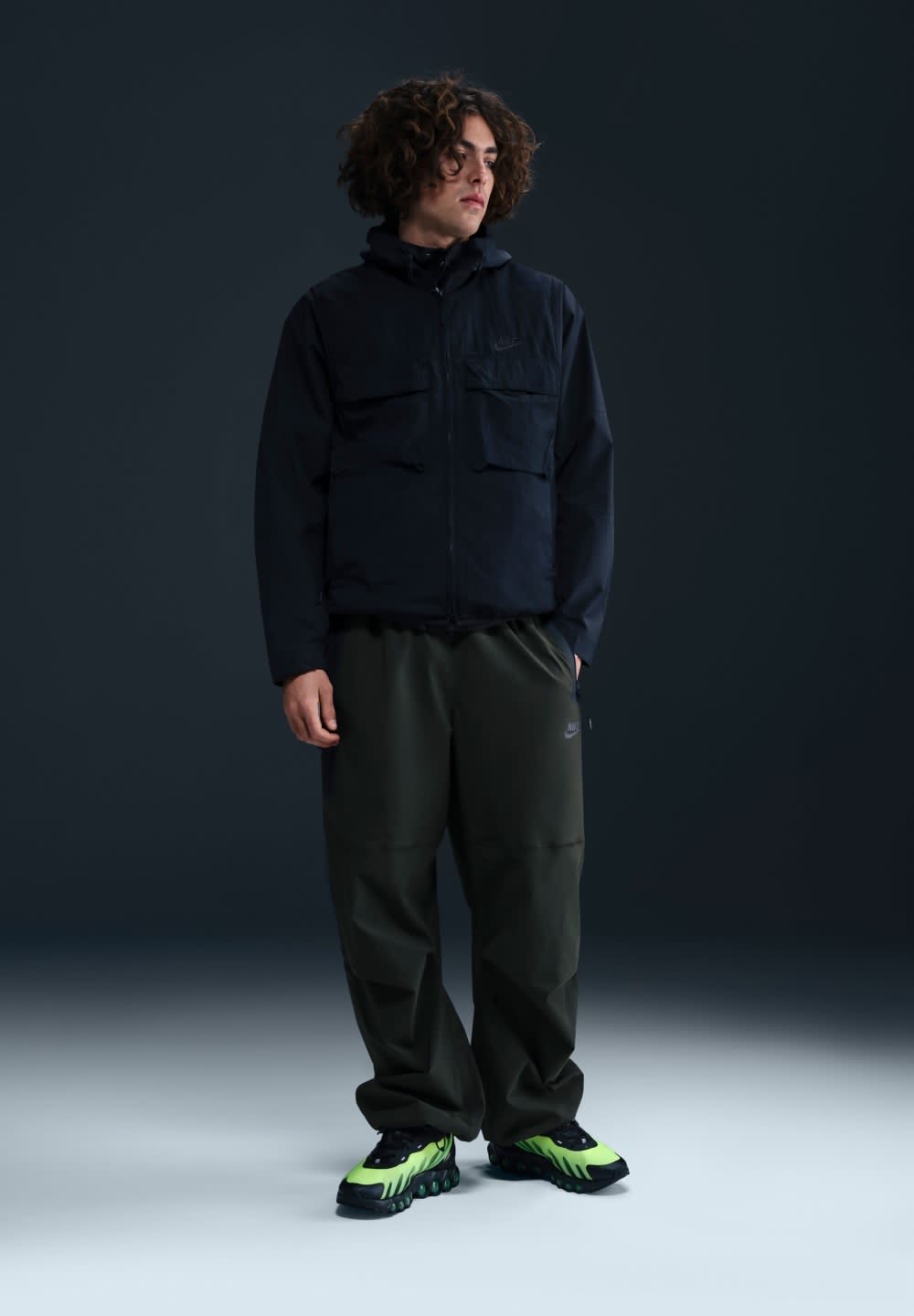 Zwarte zip-up jas met twee borstzakken, groene broek en zwarte schoenen met groene accenten. Model staat tegen een donkere achtergrond.