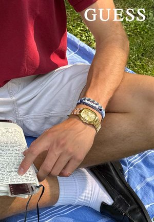 Goldfarbene achteckige Armbanduhr mit hellem Ziffernblatt und metallischem Armband, kombiniert mit blauen Perlenarmbändern, die auf einem weißen Notizbuch liegen.