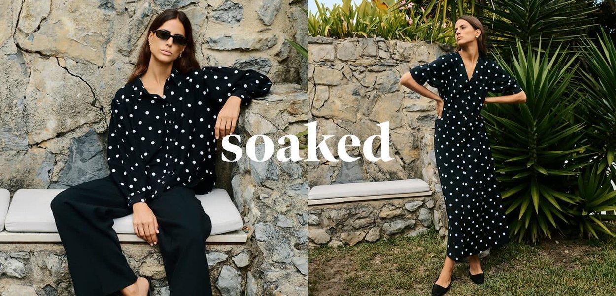 Schwarzes gepunktetes Kleid mit lockerem Schnitt, übergroßen Ärmeln und mittellanger Länge, kombiniert mit weitgeschnittenen schwarzen Hosen. Hintergrund aus Naturstein.