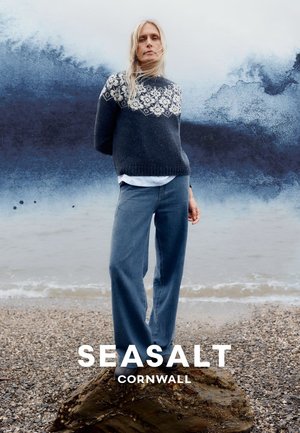 Frau steht auf einem Felsen am Strand, trägt einen blauen Pullover mit Muster und weit geschnittene Jeans, mit Meer und Himmel im Hintergrund.