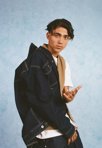 Denim jacket met een tan overlay, voorzien van grote zakken en zichtbare stiksels. Ggedragen over een wit T-shirt, tegen een lichtblauwe achtergrond.